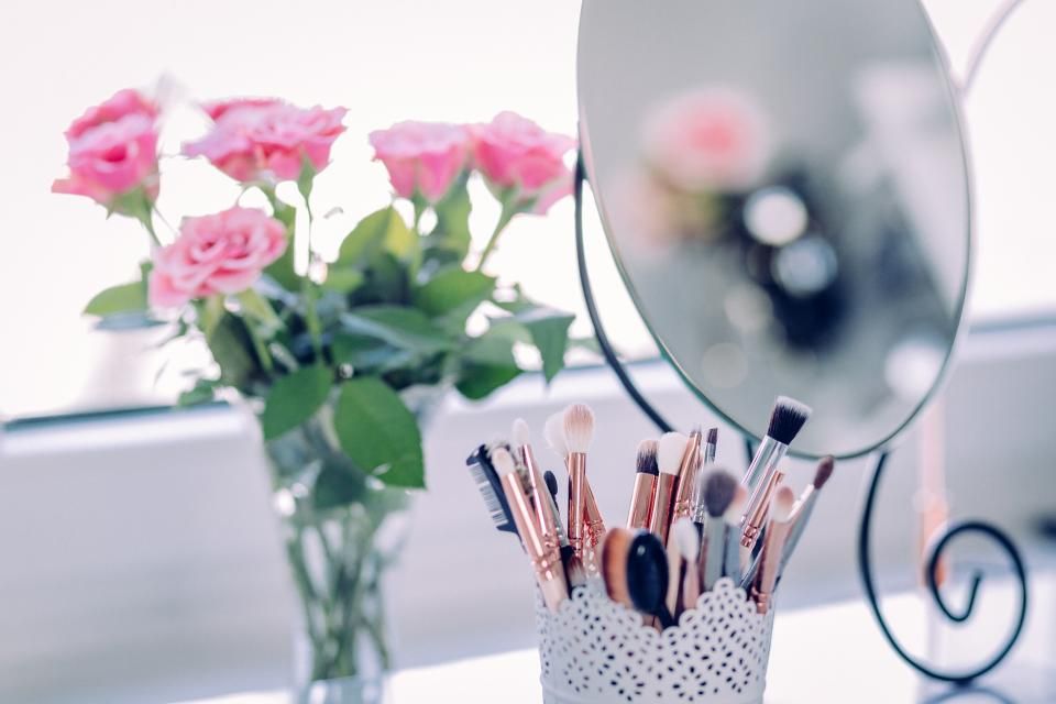 Makeup brushes in a holder next to pink roses and a small mirror on a window sill